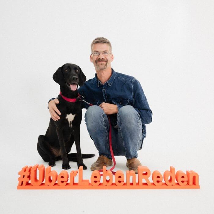 Helmut L. sitzt mit seinem Hund vor einem Aufsteller #ÜberLebenReden. Er ist Teil der gleichnamigen Aktion des neunerhauses und beantwortet Fragen zum Thema Wohnungslosigkeit. 