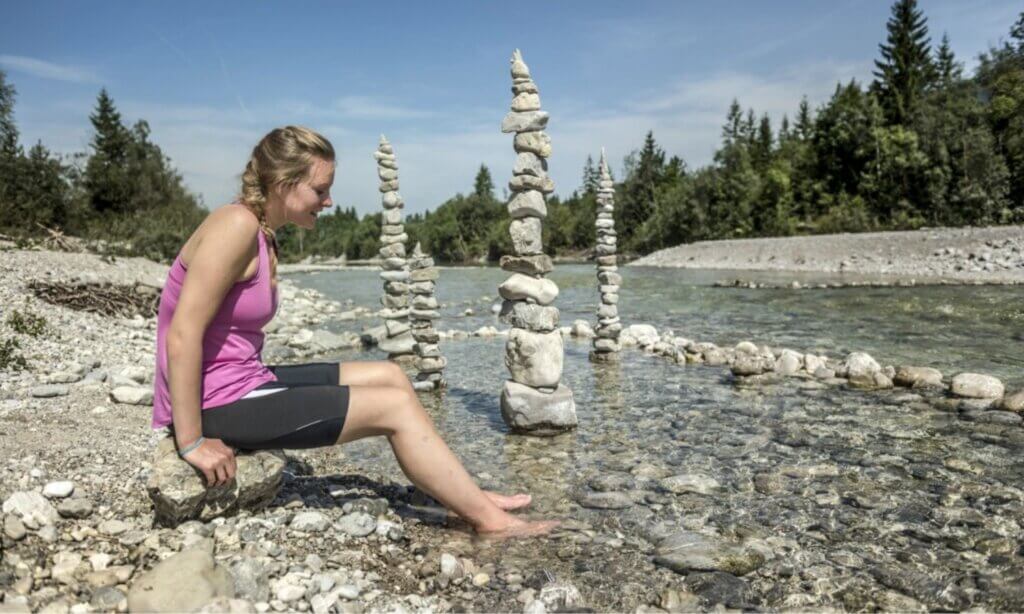 Eine Frau sitzt am Kiesstrand und hält ihre Füße ins Wasser der Isar.